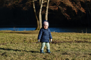 Baby girl in jacket stand in sunny spring park against river.