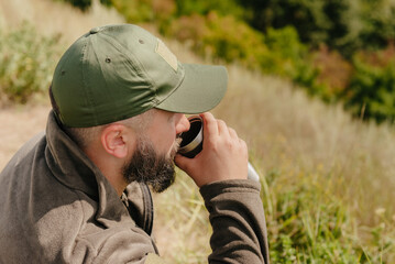 Hiking outdoors leisure concept: bearded male enjoys tea drink from metal cup while sitting outdoors in the beautiful hills natural location near river close up portrait