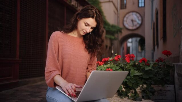 Woman typing on a laptop keyboard while sitting on decorative stones of city streets, A pretty young female using a laptop, girl student preparing for classes in nature, adult woman studying outdoors