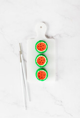 Fototapeta na wymiar Mousse cream cakes with jelly watermelon in the form of round sweet Japanese sushi, sprinkled with green coconut chips, on a serving board. Light background. Top view
