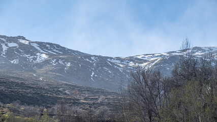 Bottom-up view to rocky mountains partly covered by snow with clear blue sky in background and empty winding paved road along clay cliff partially overgrown in the foreground.