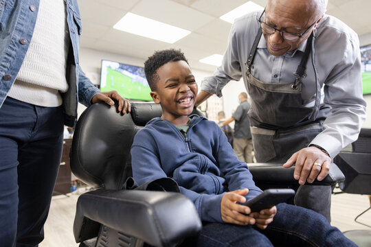 Happy boy with smart phone laughing in barber shop chair