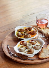 Two plates of baked snails sit on a large brown wooden plate with croutons and a glass of rose wine. Snails baked with butter and spices. Brown background. Exquisite food. French cuisine concept.