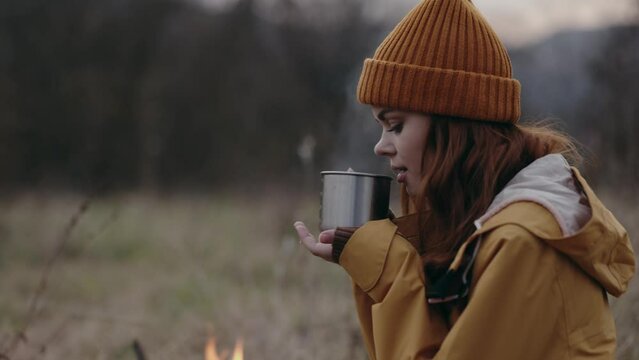A woman in a yellow hat in a camping trip drinking tea sitting by a fire in the countryside and boiling water in an herbal tea kettle in a camping mug to keep warm in the fall on a trip