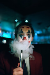 Guy in suit and tie with the Joker makeup puffs smoke out of his mouth. Autumn holiday. All Saints' Night. Halloween party in nightclub with decorations and scary costumes.