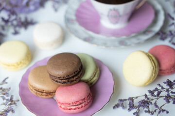 Mix of appetizing macarons and delicious coffee in porcelain cup on white table decorated with blurred flowers. Dry flowers. Greeting card.