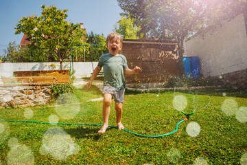 Laughing handsome boy run around water sprinkler in the garden - obrazy, fototapety, plakaty
