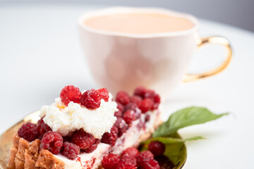 Delicious piece of cake with fresh raspberries and blurred cup of aromatic coffee in background. Blurred white background. Sweet concept. Excellent image for dessert banners and advertisements.