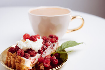Delicious piece of cake with fresh raspberries and blurred cup of aromatic coffee in background. Blurred white background. Sweet concept. Excellent image for dessert banners and advertisements.