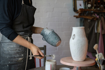 Woman in apron works with pottery standing near round wooden table with equipment. Professional...