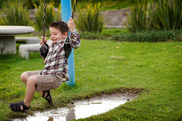 Niño feliz en el parque en el columpio jugando divertido colgado arriba de un charco de agua lleno de lodo al aire libre disfrutando de un hermoso día - obrazy, fototapety, plakaty