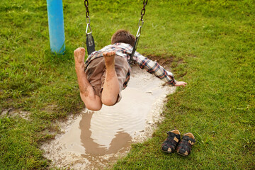 Niño feliz en el parque en el columpio jugando divertido colgado arriba de un charco de agua lleno de lodo al aire libre disfrutando de un hermoso día - obrazy, fototapety, plakaty