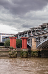 Naklejka premium London, England, UK - July 6, 2022: From Thames River. Oxblood red pillars protecting Blackfriars railway bridge NW shore under thick gray cloudscape.