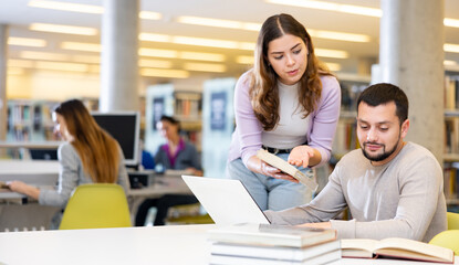 Positive man and woman spending time together in library, talking, work on laptop