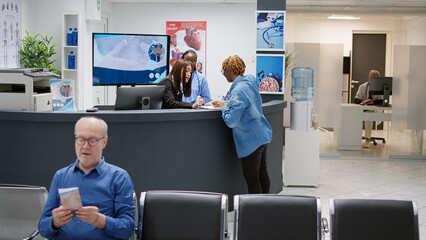 African american patient filling in checkup report at reception counter, receiving help from asian receptionist. Woman having medical consultation appointment, waiting area lobby.