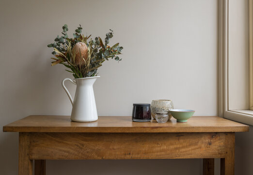 Australian native flowers in white jug wtih little bowls on oak side table against beige wall