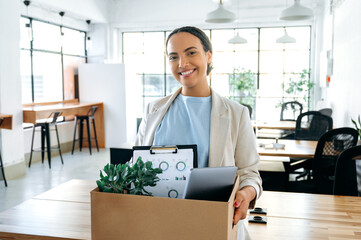 Happy young brazilian or hispanic female employee, holding cardboard box, standing in modern office...