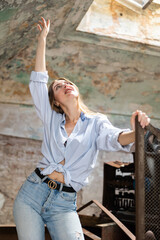 Young desirable brown haired woman wearing shirt, posing in derelict building raising her hands.