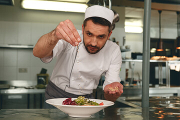 Chef sprinkles salt to salad of fresh vegetables in kitchen of restaurant. Tasty and healthy food