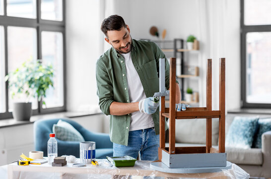 renovation, diy and home improvement concept - happy smiling man in gloves with paint roller painting old wooden table in grey color