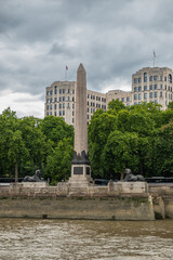 London, England, UK - July 6, 2022: From Thames River. Cleopatra's Needle statue, backed by green foliage belt and high rise buildings under gray cloudscape.