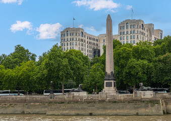 London, England, UK - July 6, 2022: From Thames River. Cleopatra's Needle statue, backed by green foliage belt and high rise building under blue cloudscape.