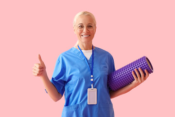 Mature physiotherapist with foam roller showing thumb-up on pink background