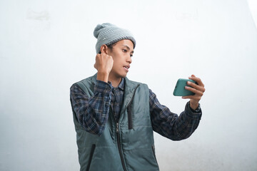 Asian young man wearing dark blue vest and beanie, isolated on white background looks happy looking at his phone screen.