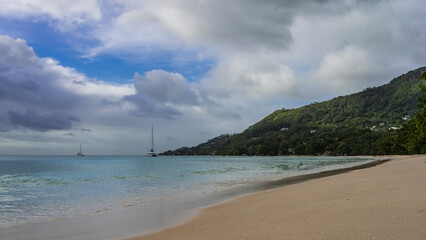 Fototapeta na wymiar Sandy beach on a tropical island. The turquoise ocean is calm. Yachts are visible in the distance. A green hill against a background of blue sky and clouds. Seychelles. Mahe. Beau Vallon