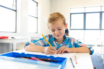 Smiling caucasian elementary schoolboy writing on book while sitting at desk in school