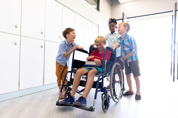 Cheerful multiracial schoolboys standing by male classmate sitting on wheelchair in school corridor