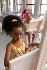 Obraz premium Biracial elementary schoolgirl holding paint palette while painting on easel during drawing class