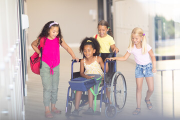 Smiling multiracial elementary schoolgirls assisting female biracial classmate sitting on wheelchair