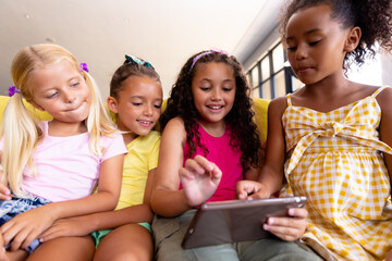 Smiling multiracial elementary schoolgirls using digital tablet while sitting on couch in school