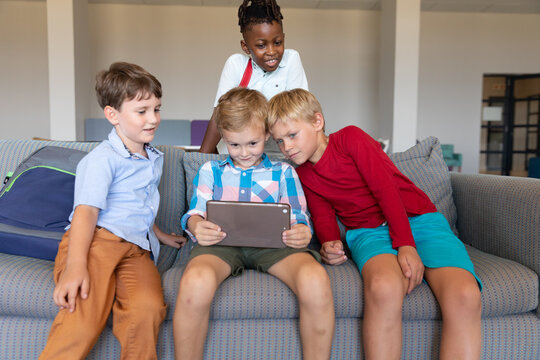 Multiracial elementary schoolboys looking at digital tablet while sitting on couch in school