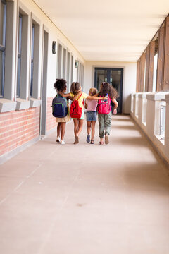 Full length of multiracial elementary schoolgirls with backpacks and arm around walking in corridor