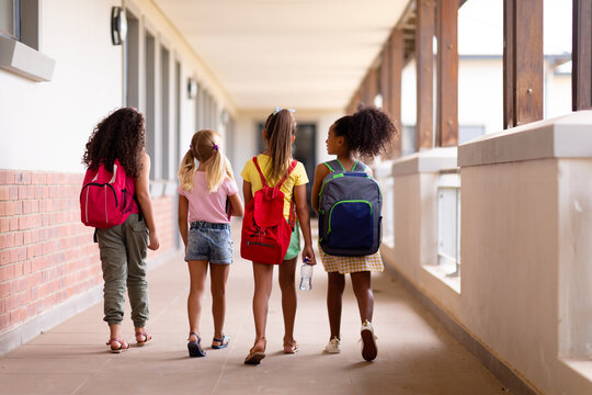 Full length rear view of multiracial elementary schoolgirls with backpacks walking in corridor