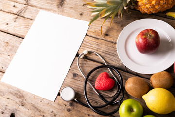 High angle view of various fruits with stethoscope and heart shape by blank paper on wooden table