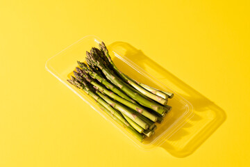 High angle view of raw asparagus in plastic container with shadow by copy space on yellow background