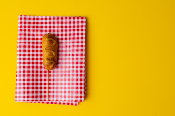 Overhead view of corn dog with mustard sauce on checked pattern napkin over yellow background