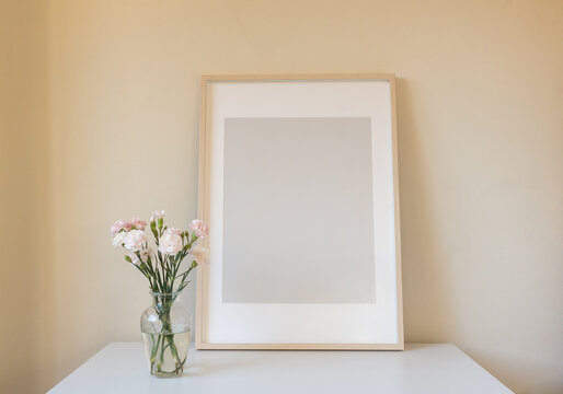 Pink carnations in glass vase with large blank rectangular picture frame on white table against beige wall (selective focus)