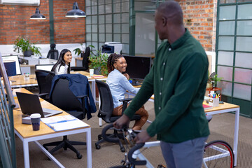 African american young businessman with bicycle looking at happy multiracial young female colleagues
