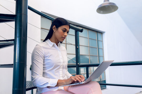 Biracial young businesswoman using laptop while sitting on steps in modern office