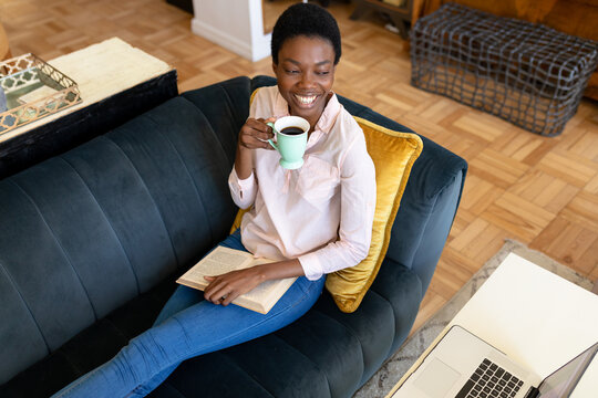 Happy african american woman sitting with coffee cup and book on sofa in living room