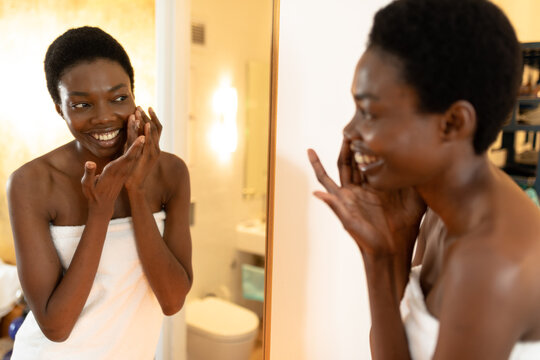 Smiling african american woman applying cream while looking at mirror reflection in bathroom
