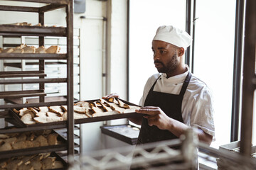 African american mid adult male baker removing baked bread tray from rack in bakery