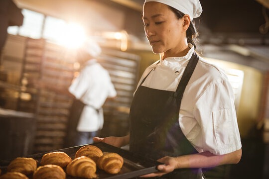 Asian mid adult female baker holding baking tray with croissant while working in bakery