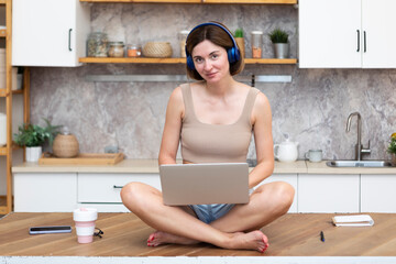Young beautiful woman in headphones studying online at home. She sits on the table in the kitchen	