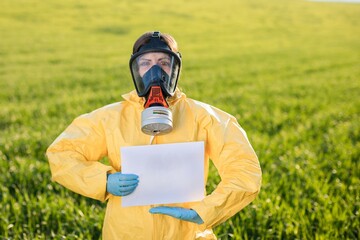 Person in overalls and gas mask holds holds a sheet of paper with the exclamation Save the Planet while standing on green field on sunset.