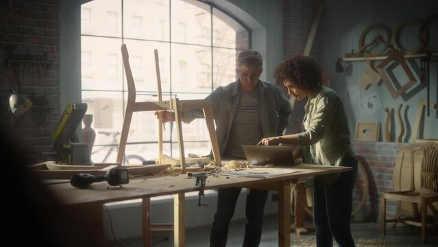 Small Business Owners of a Furniture Workshop Using Laptop Computer and Discussing the Design of a New Wooden Chair. Middle Age Carpenter and a Young Female Apprentice Working in Loft Studio.